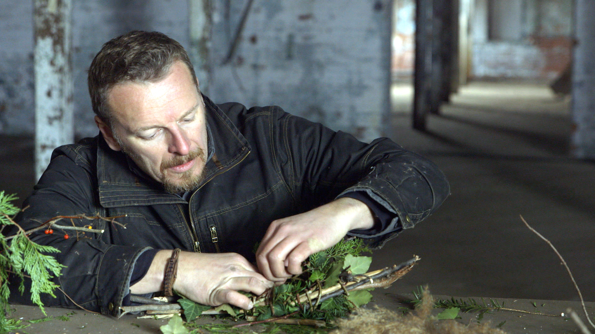 A man in a black jacket is sitting at a table, focusing intently on arranging branches and leaves to mimic animal homes. The background shows a rustic, open space with exposed brick and wooden beams.