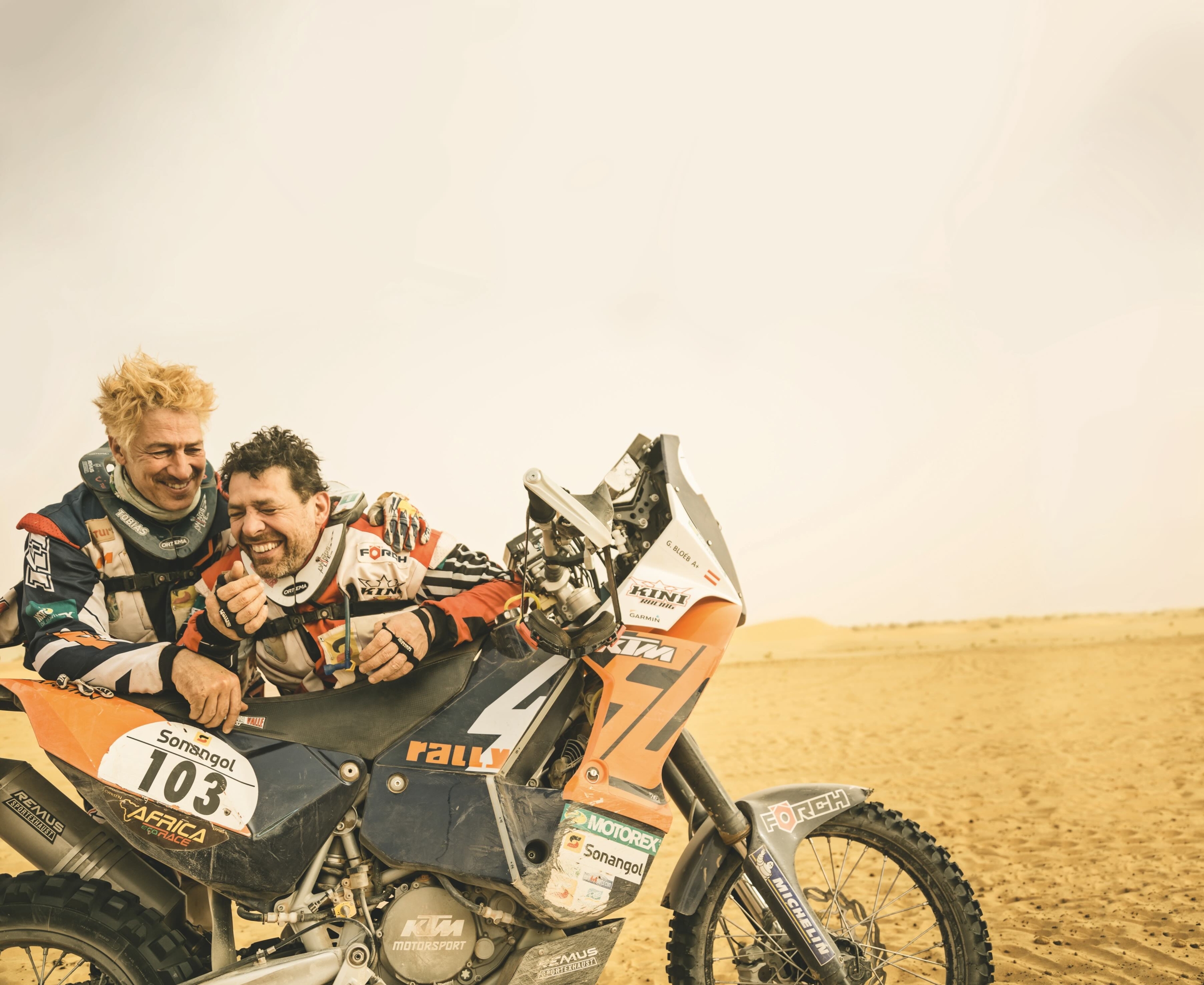 Two men in racing gear happily lean on a dirt bike in a desert setting. One bike shows the number 103 and the other 47. The sky is overcast, and the landscape consists of sand dunes in the background.