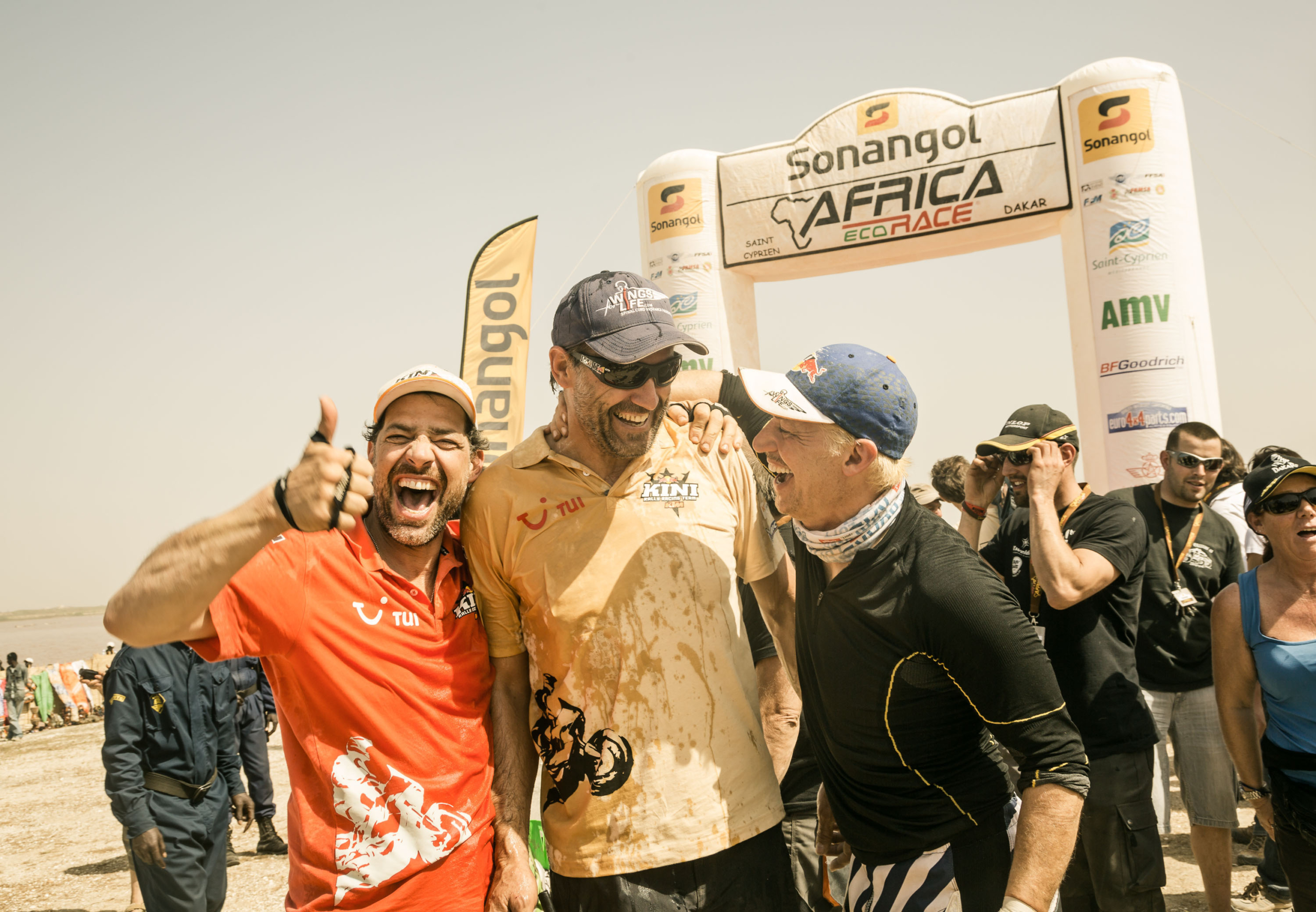 Three men celebrating at a race finish line, smiling broadly and embracing. They are standing in front of a large inflatable arch that reads Africa Eco Race with various logos. The atmosphere is joyful and crowded with spectators in the background.