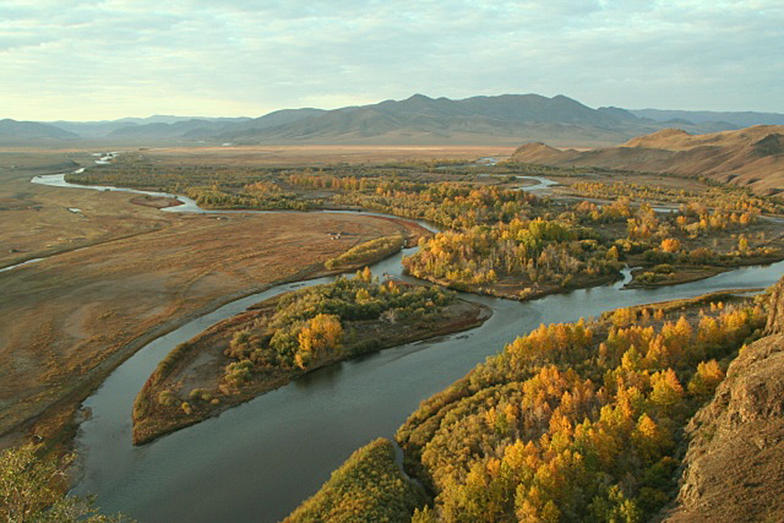 A winding river, reminiscent of the Amur, flows through a vast landscape with autumn-colored trees and grassy plains. The distant mountains rise under a partly cloudy sky, creating a serene and expansive view.