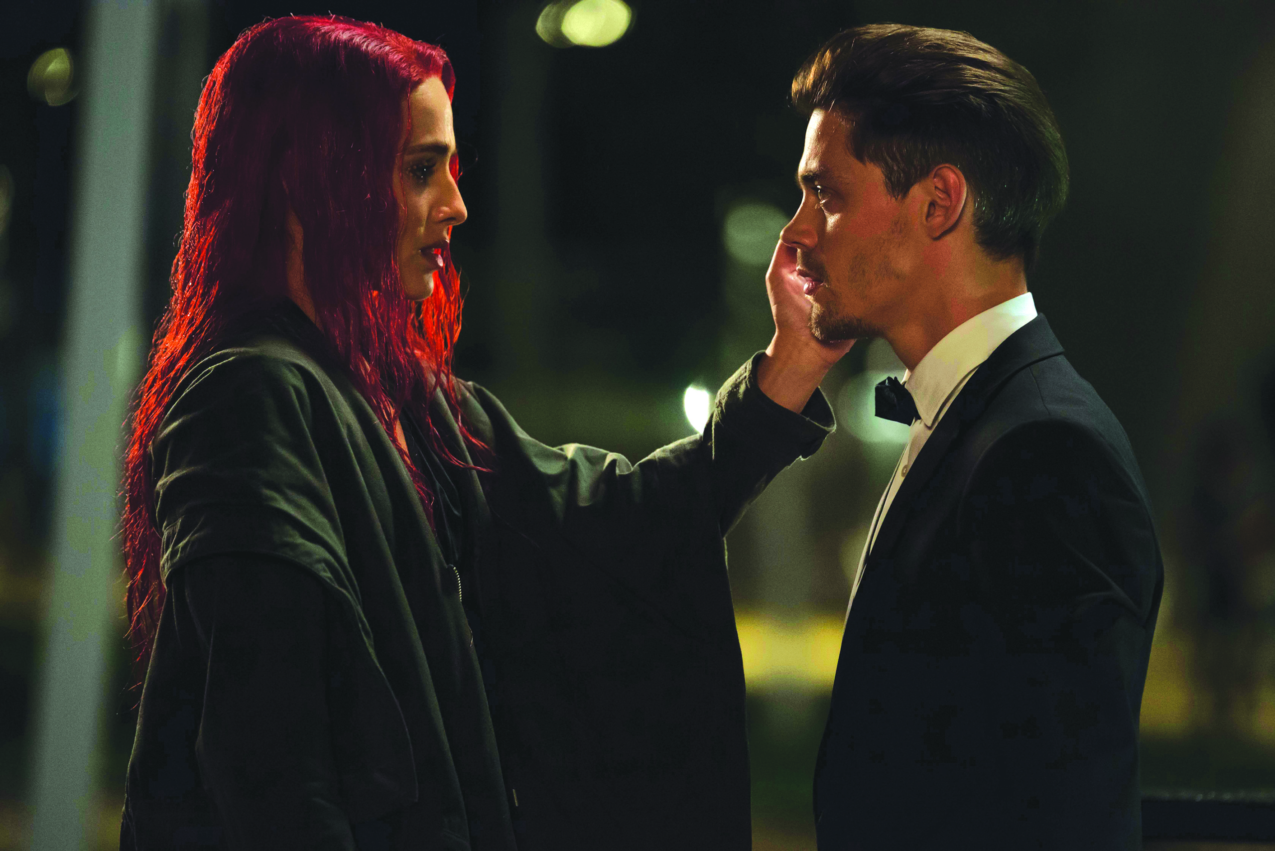 A woman with vibrant red hair gently touches the face of a man in a tuxedo. They are standing close together, exchanging a meaningful look in a dimly lit setting. The atmosphere suggests a moment of emotional connection.