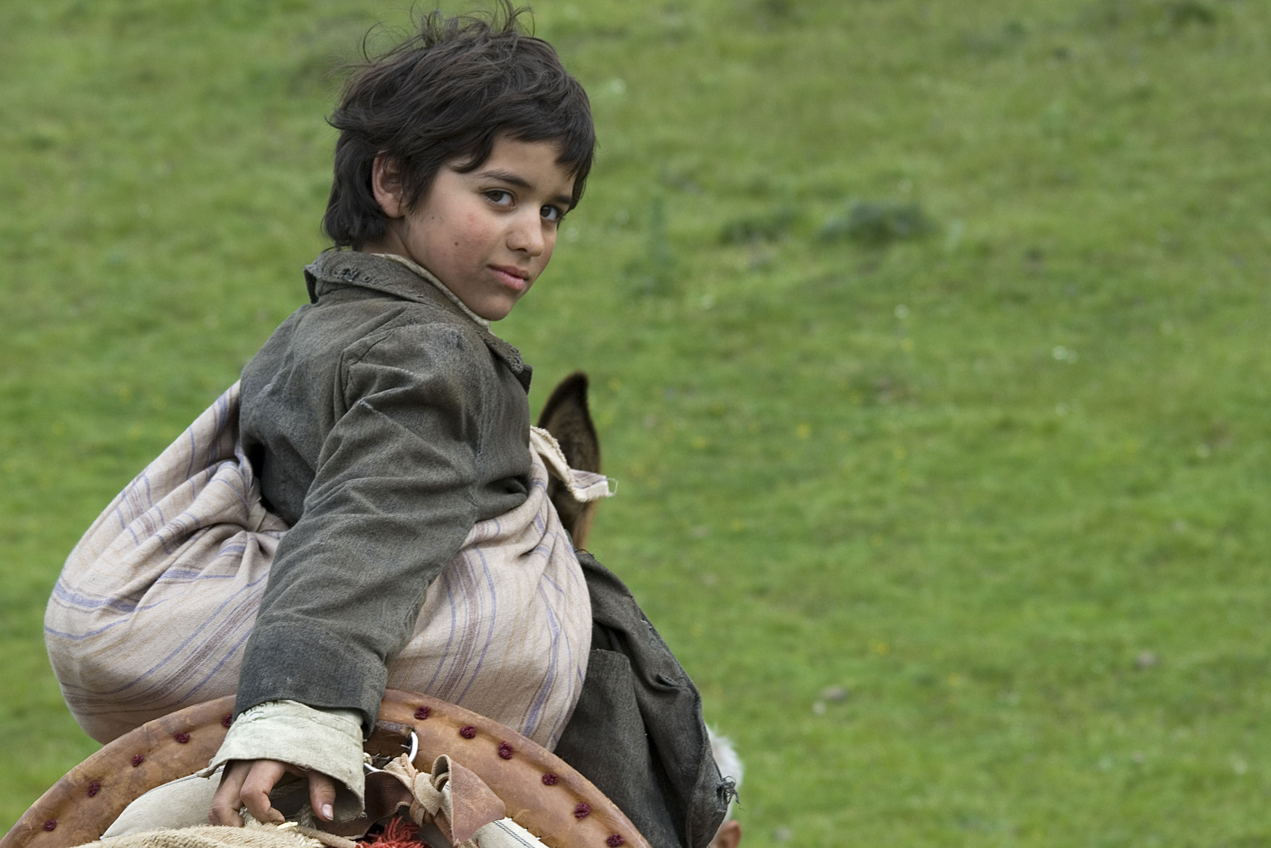 A young child with tousled dark hair and a weathered coat sits on a pack animal against a green grassy backdrop, much like one who has wandered among wolves. The child looks over their shoulder with a curious expression.