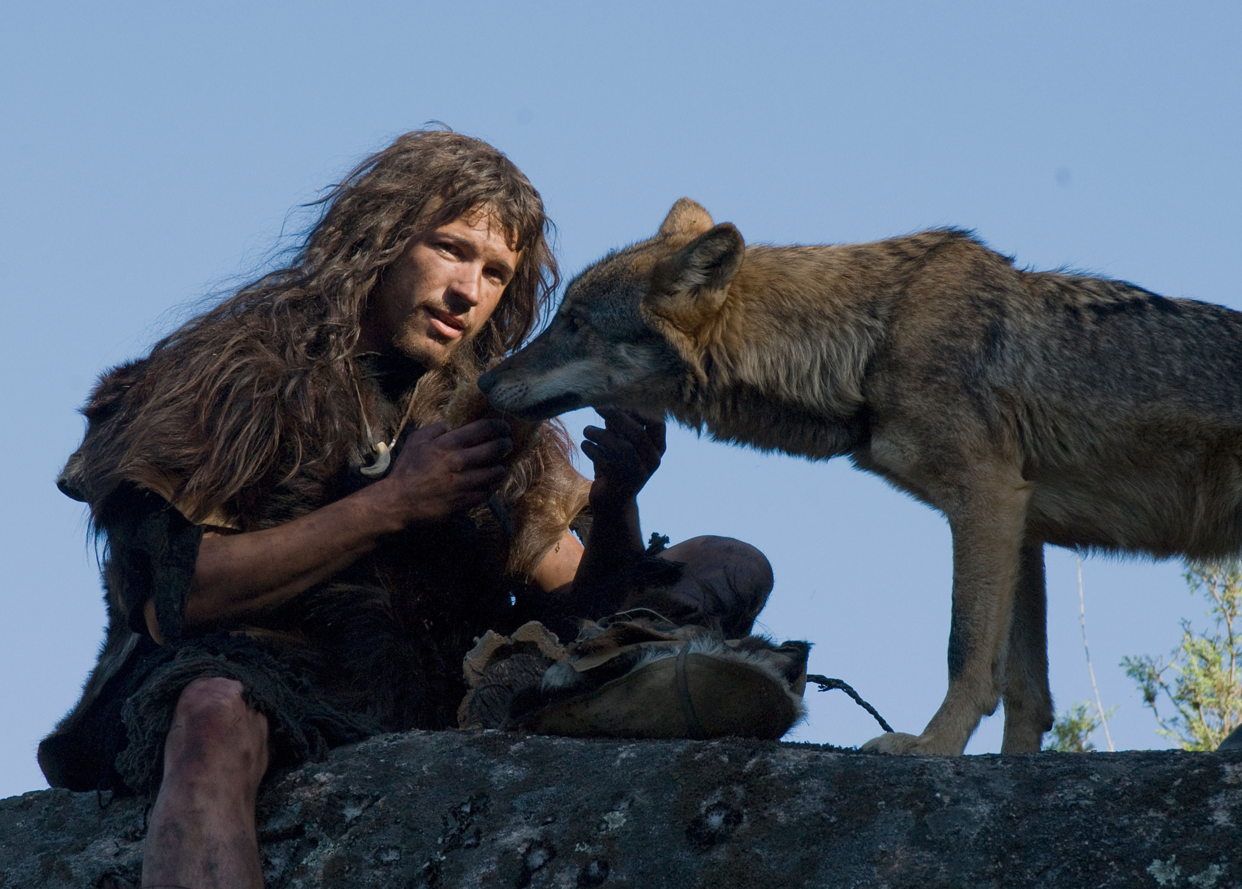 A person in rustic clothing with long hair sits on a rock, engaging closely with a wolf among wolves. The scene is set outdoors against a clear blue sky, and the wolf appears attentive and engaged with the person.