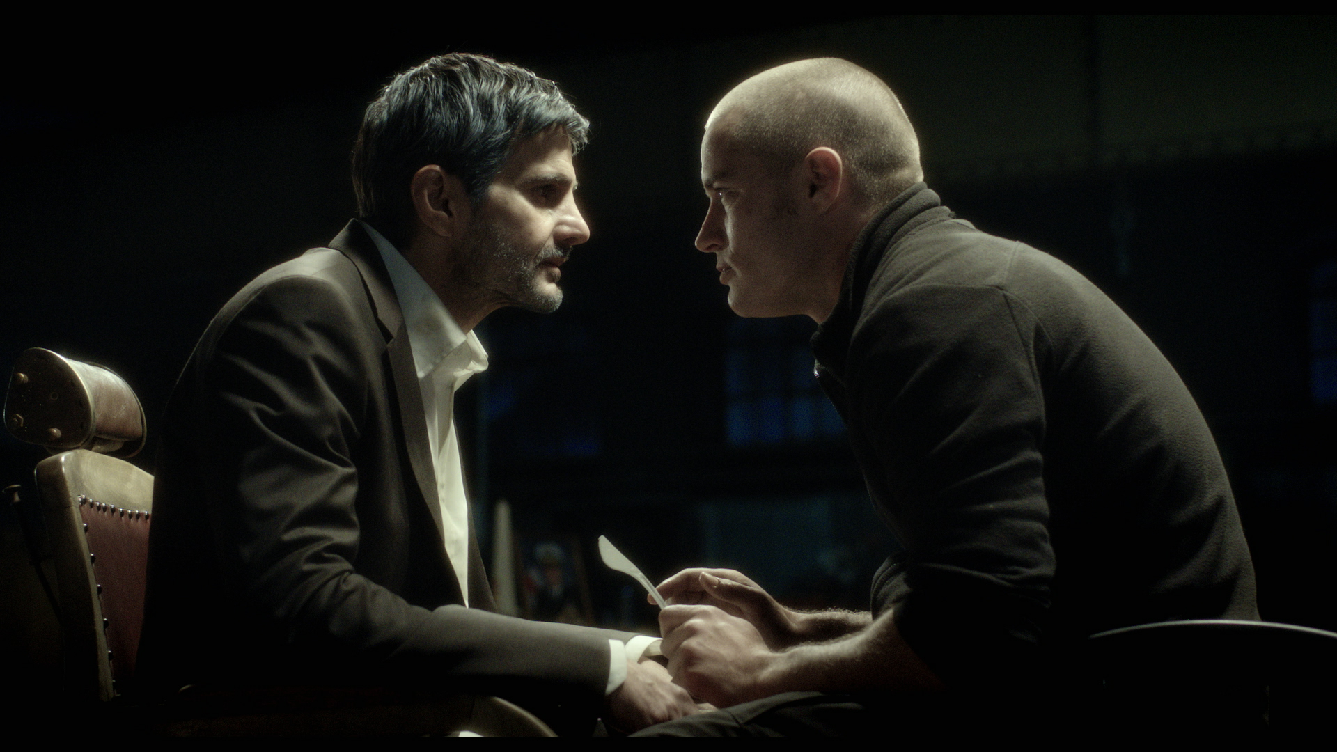 Two men sitting face-to-face in a dimly-lit room, both with serious expressions. The man on the left has dark hair and a beard, while the man on the right is bald. The setting conveys tension and confrontation.