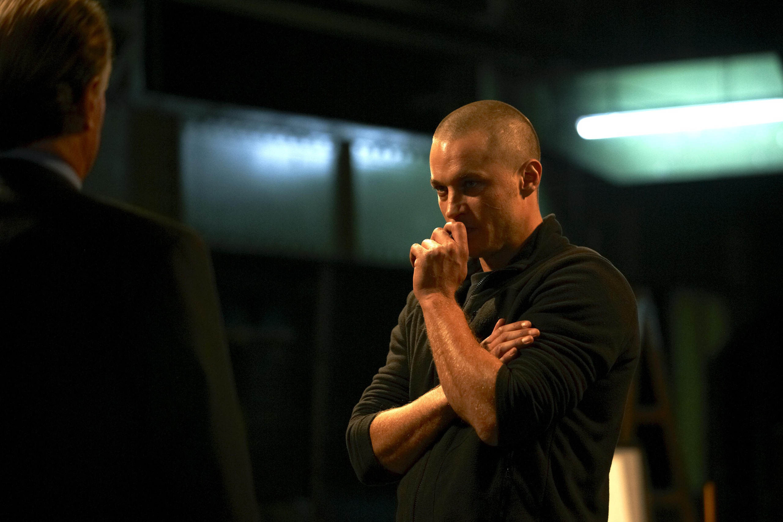 A man with a serious expression stands in a dimly lit room, one hand raised to his chin and the other crossed over his chest. Another person is partially visible, facing him. A fluorescent light is visible in the background.