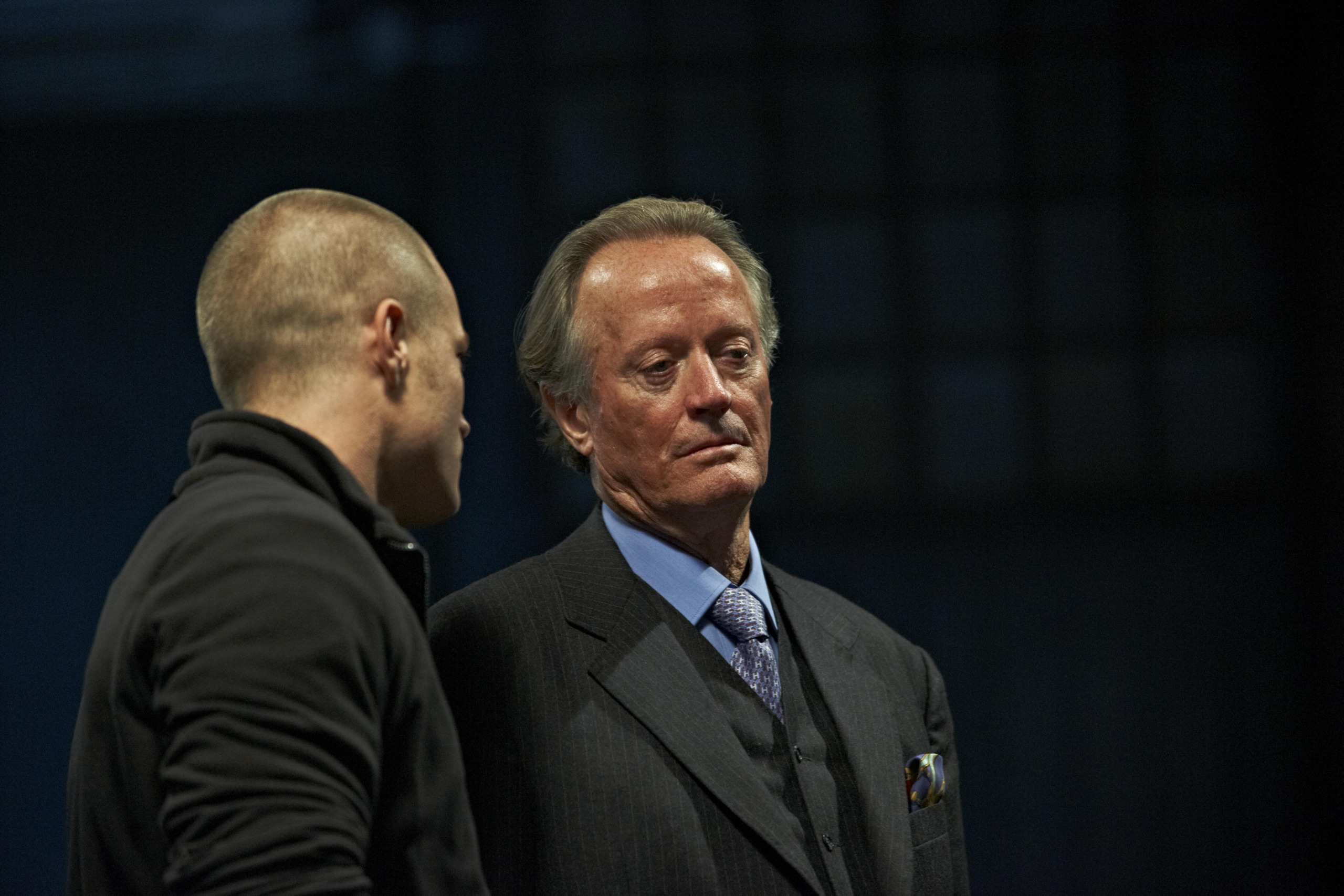 Two men are in conversation in a dimly lit area. The man on the left is bald and wearing a black jacket, facing away. The man on the right, with gray hair, is wearing a formal suit and looking ahead thoughtfully.