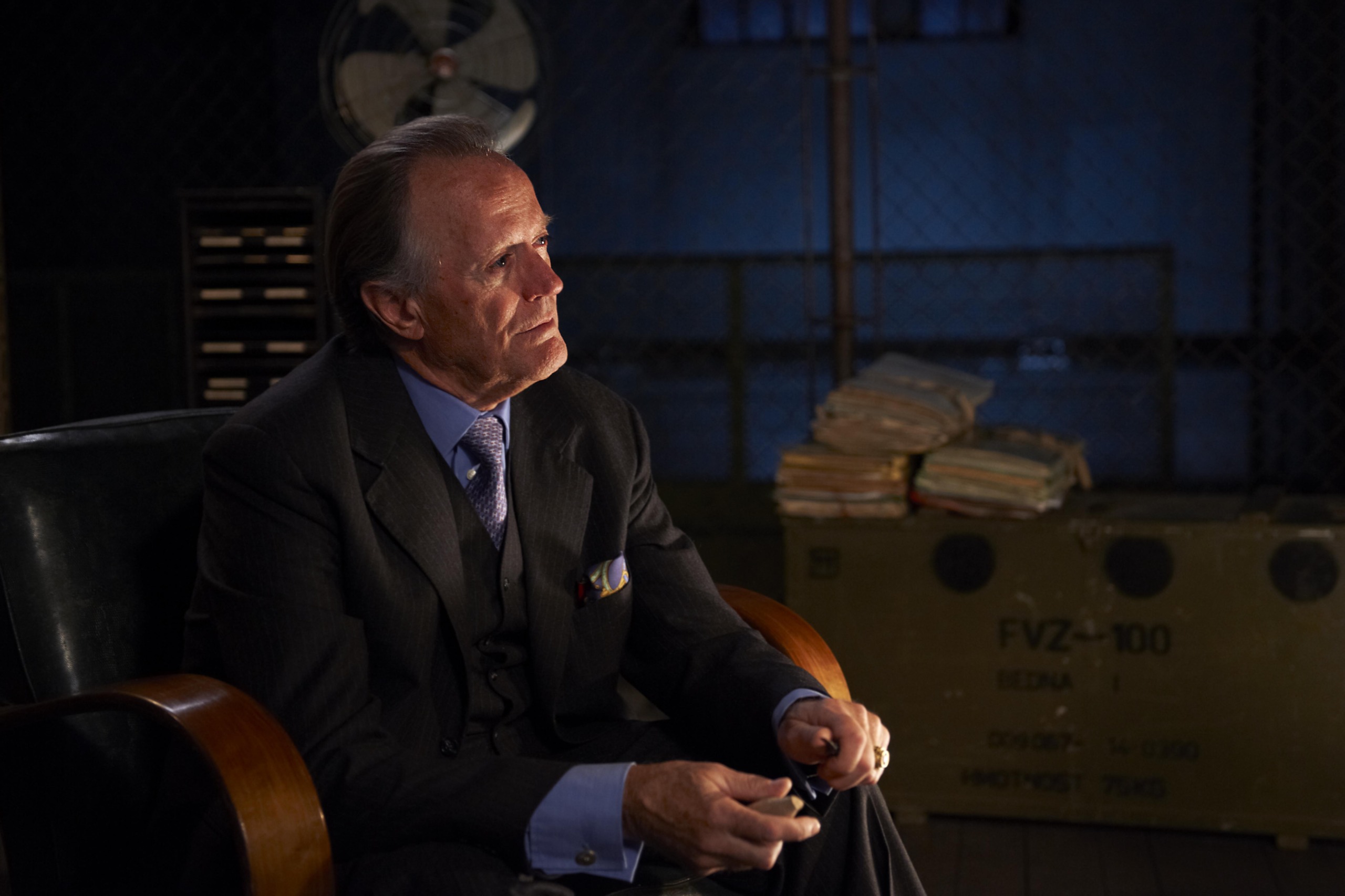 An older man in a suit sits in a dimly lit room with a serious expression. He holds an object in his hands. Behind him are stacks of folders and a large metal container. A fan and chain-link fence are visible in the background.