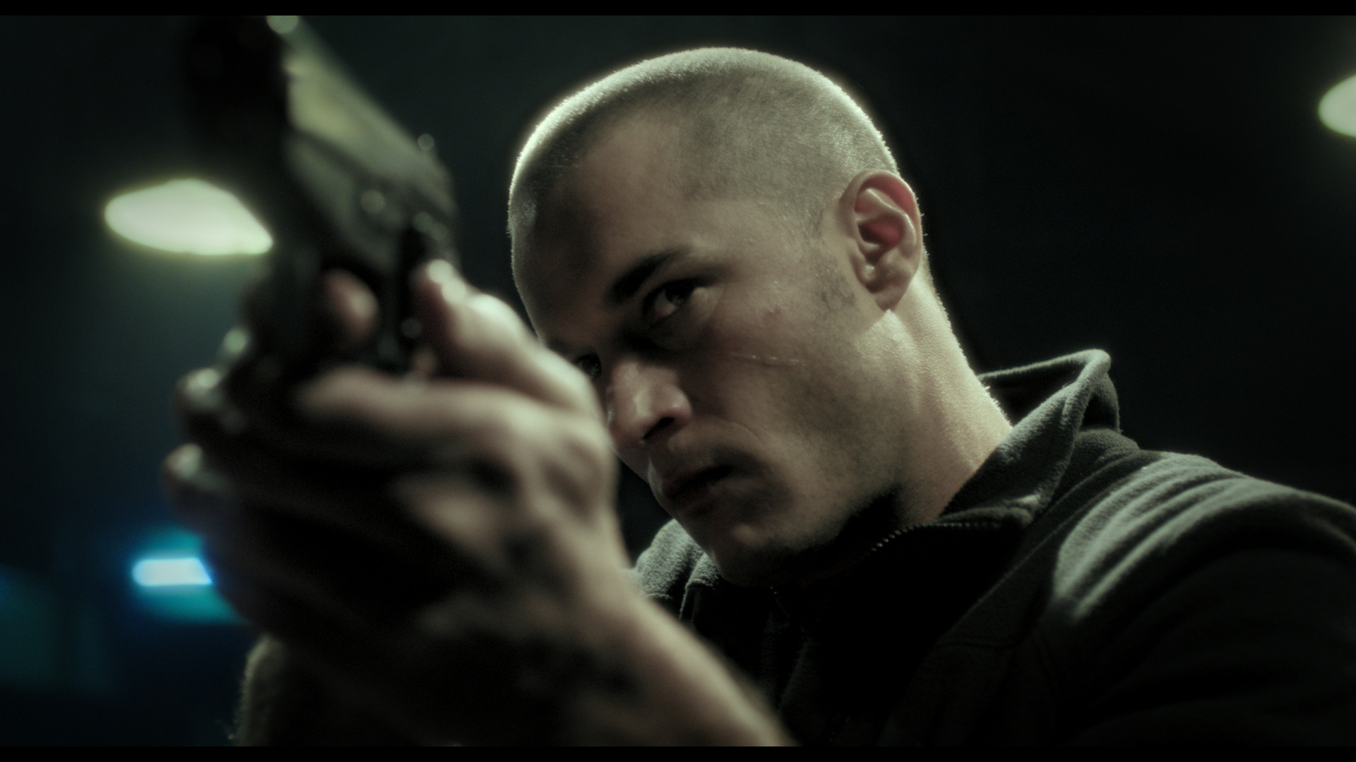 A person with a shaved head aims a handgun with a serious expression, under dim lighting. The background is blurred, focusing on the individuals intense look and the gun.