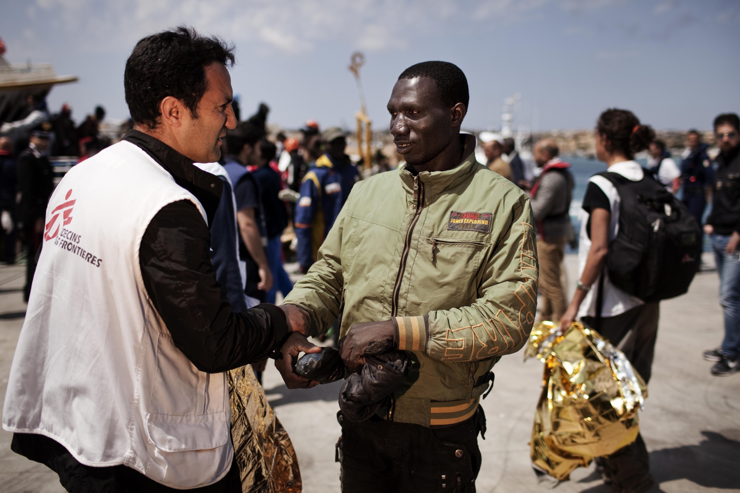 A humanitarian worker from Doctors Without Borders, wearing a white vest with a red logo, shakes hands with a man in a green jacket. They stand outdoors near a bustling port, where people and boats move in the supportive and friendly atmosphere.