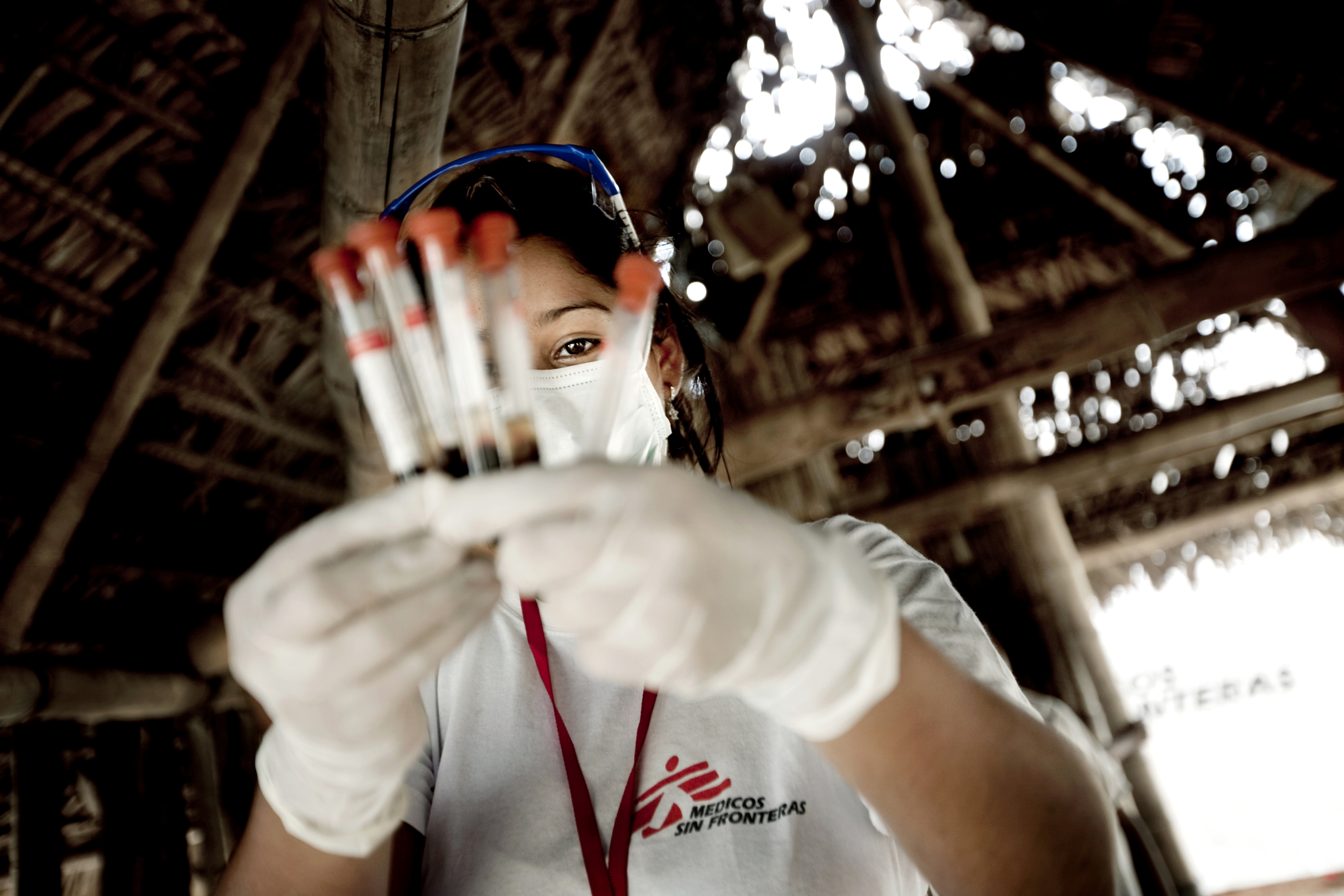 A healthcare worker in protective gear, under a thatched roof, cradles several vials of blood samples. Dressed in a white shirt emblazoned with the logo of Doctors Without Borders, they exemplify dedication and courage on the front lines.