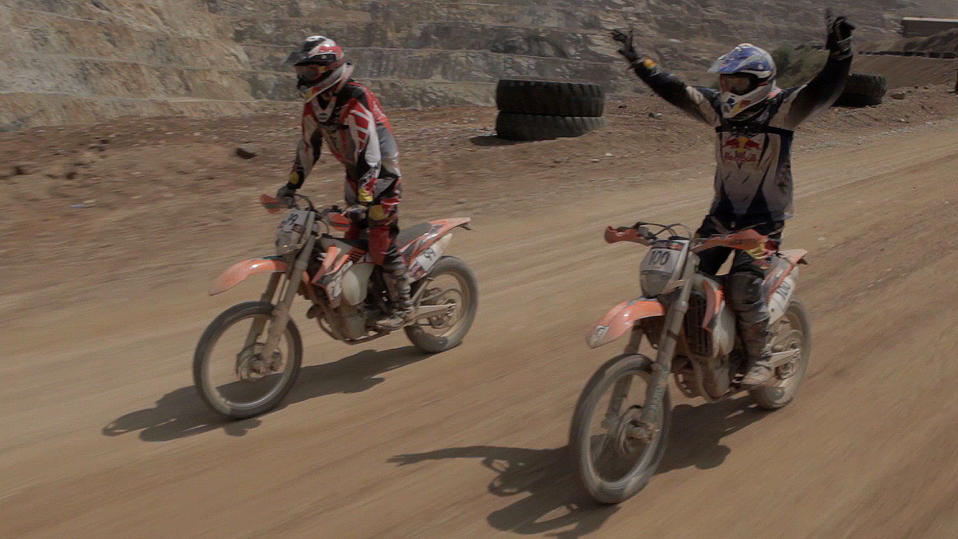 Two motorcyclists on dirt bikes ride on a dusty track. The motorcyclist on the right raises both arms in celebration, while the other maintains a steady grip on the handlebars. Both wear protective gear and helmets. Rocky terrain is visible in the background.
