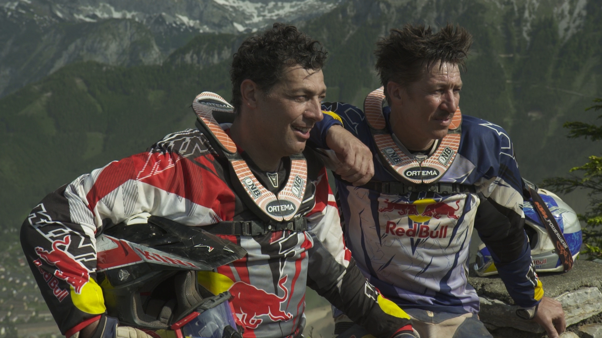 Two motocross riders in protective gear stand on a mountain overlooking a valley. They rest an arm around each others shoulders, with helmets nearby. Snow-capped peaks and dense forests form the scenic backdrop under a clear sky.