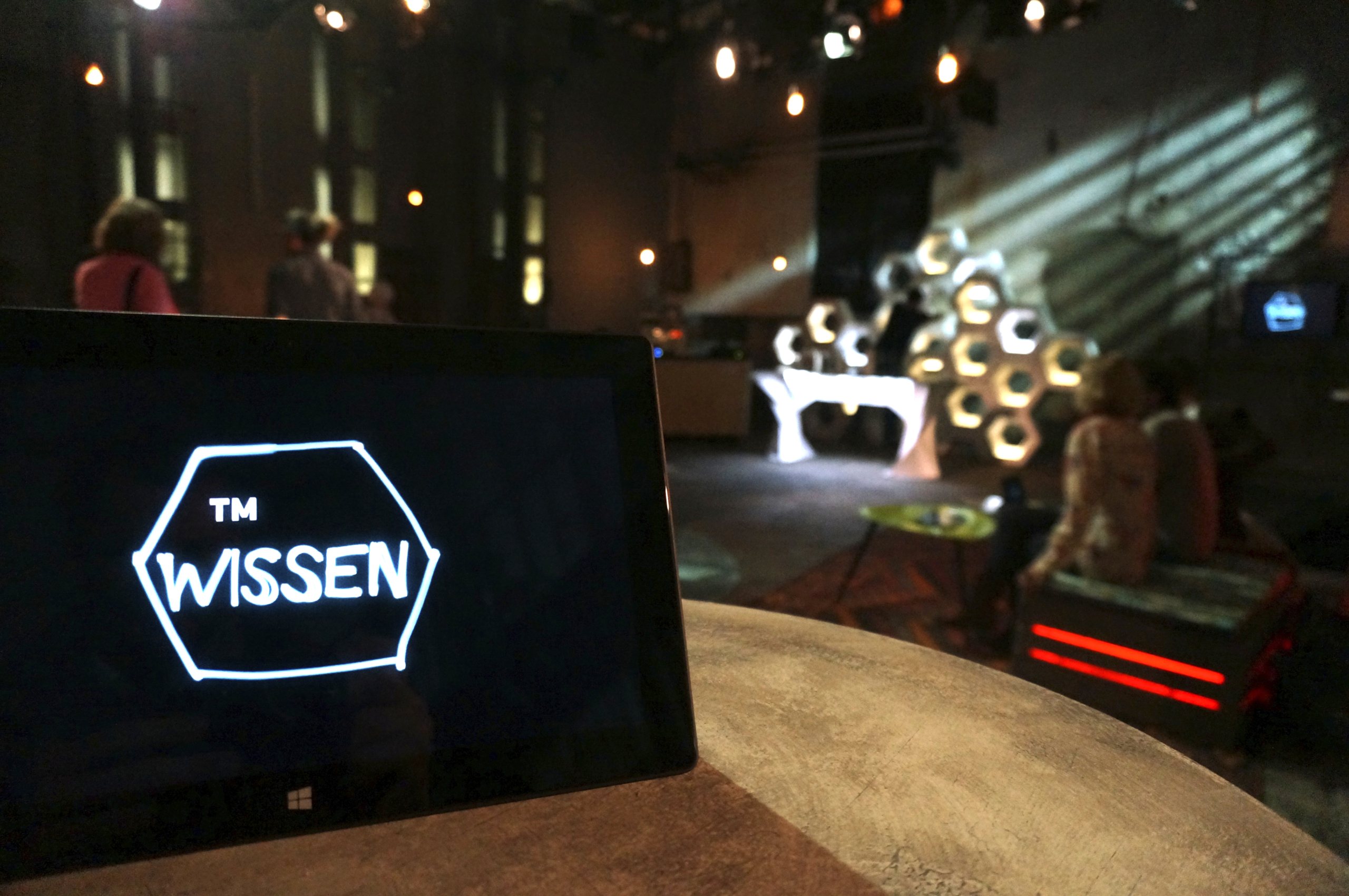 A dimly lit room with a focus on a tablet displaying WISSEN. In the background, a modern setup with circular light fixtures and silhouettes of people, creating an artistic and ambient atmosphere.