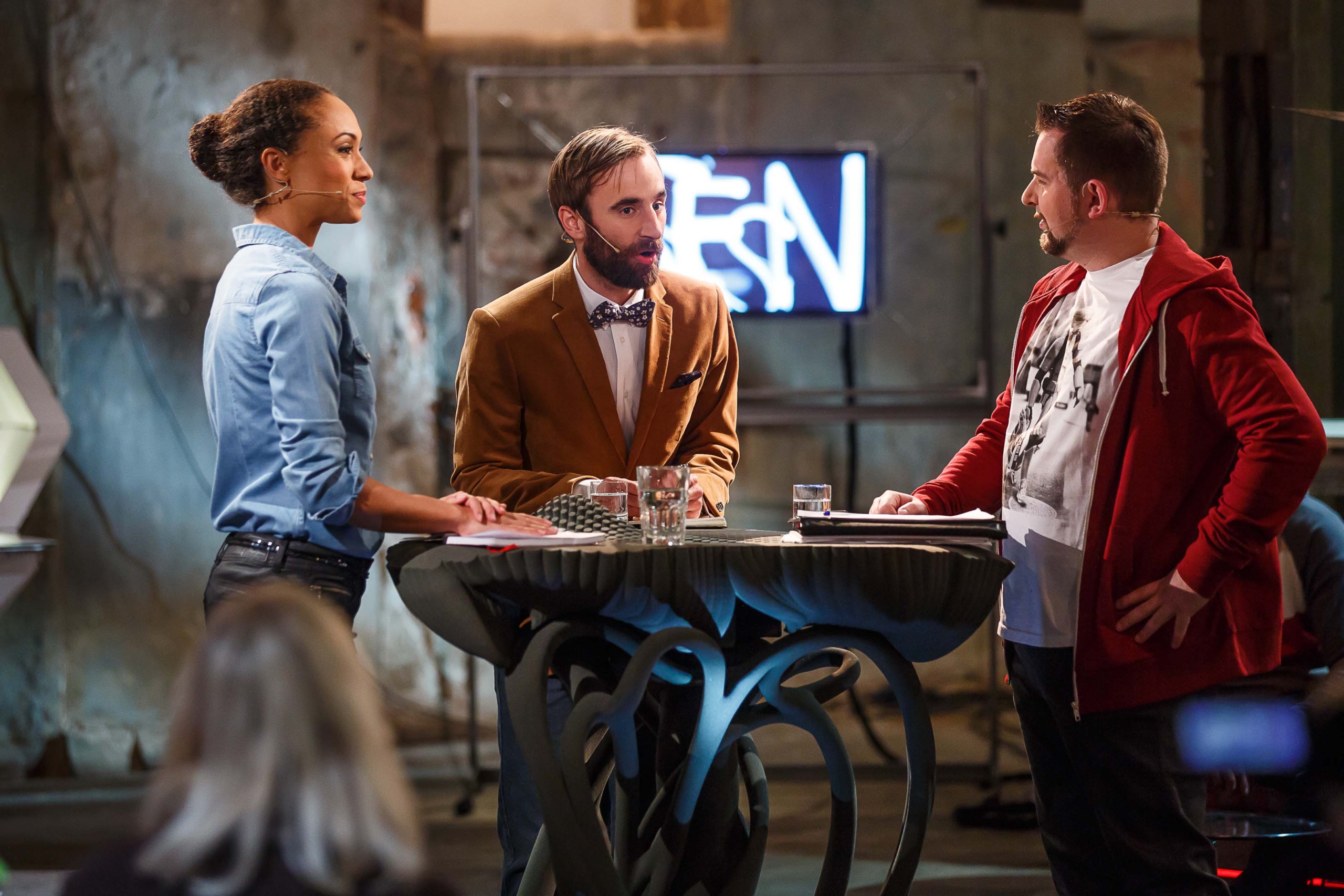 Three people are having a discussion around a round table in an indoor setting. A neon sign is in the background. One person wears a brown jacket, another a blue shirt, and the third a red hoodie. Glasses and papers are on the table.