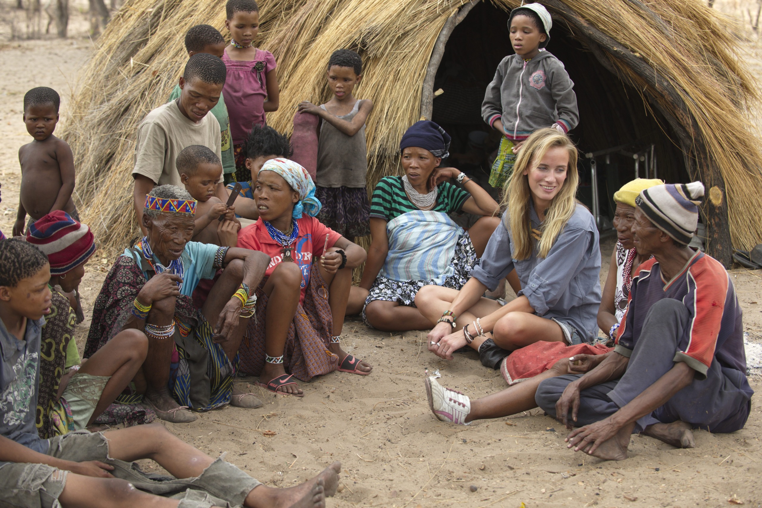 A group of Bushmen sits outside a thatched shelter in a sandy area, their vibrant clothing creating a vivid mosaic. Amidst them, a young woman with blonde hair smiles like an elegant model, engaged in lively conversation.