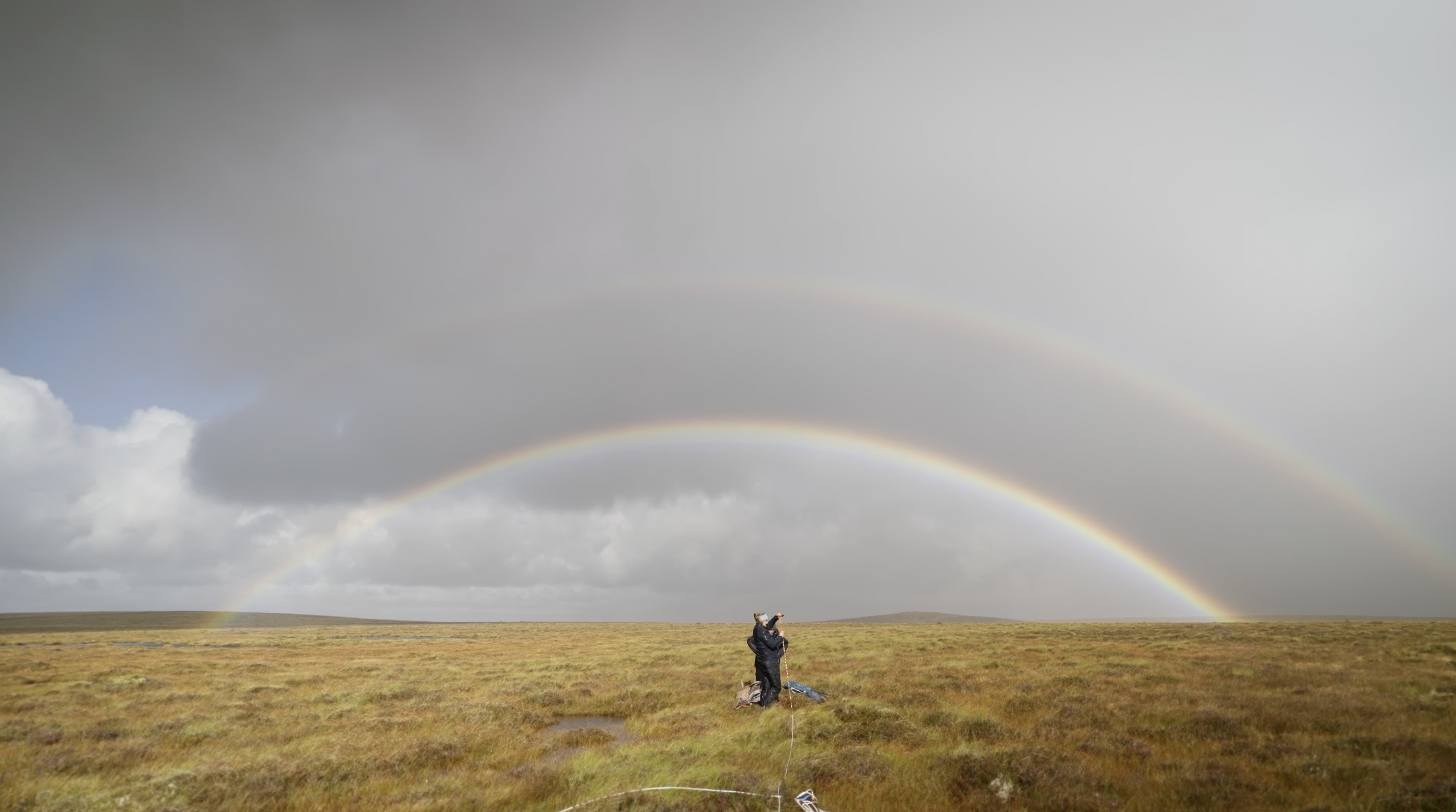A person stands in an open grassy field under a cloudy sky, gazing at a vivid double rainbow arching across the horizon—an awe-inspiring scene that evokes the wonder of discovering a New World. The landscape is flat and expansive, with muted yellow and brown tones.