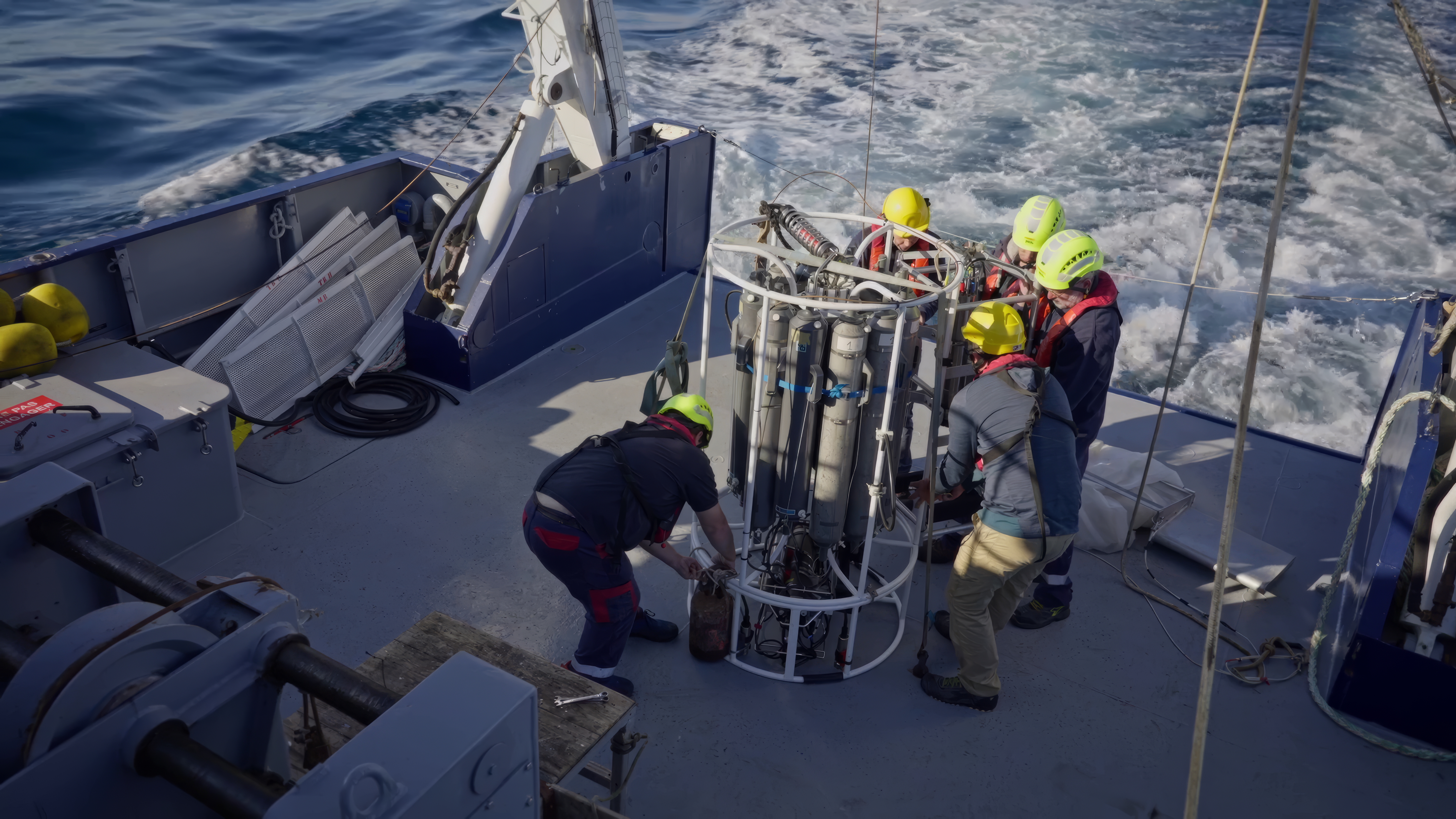 Four people in safety gear on a research vessel’s deck work together to handle a large scientific instrument surrounded by metal bars, collecting data as they explore the ocean waves of the New World behind the boat.