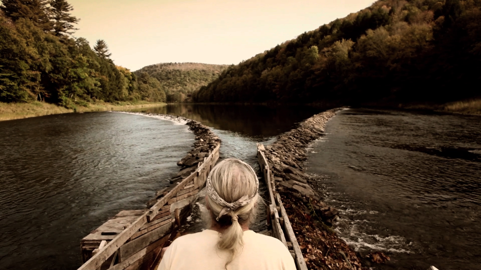 A person with long gray hair stands at the edge of a river, contemplating an eel weaving through the water by a V-shaped stone dam. Trees line both sides under a clear sky, and distant hills are visible in the background.