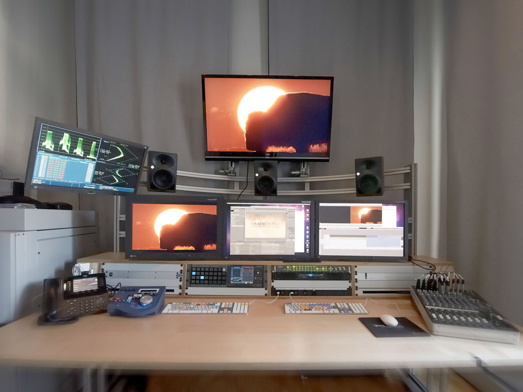 A video editing workstation featuring multiple monitors displaying a sunset scene, speakers, a mixing console, and various audio-visual equipment. A dimly lit room with a focus on the editing process.