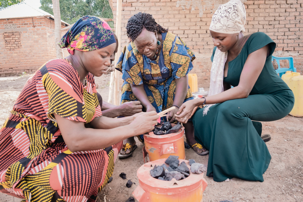 Three women in colorful clothing sit around a coal stove, engaging in a green business initiative. Focused and collaborative, they work together in a rural setting, surrounded by a brick wall and tools.