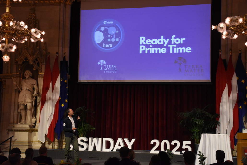 A man stands on stage presenting in front of a large screen displaying Ready for Prime Time at SWDays25. Flags, chandeliers, and classical statues decorate the ornate room, blending tradition with the innovations of the Digital World.