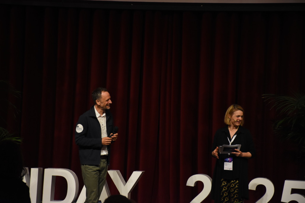 Two people stand on a stage with a red curtain backdrop, one holding a microphone and the other holding a tablet. The large white letters and numbers reading SWDays25 are partially visible behind them, highlighting the events role in the digital world.