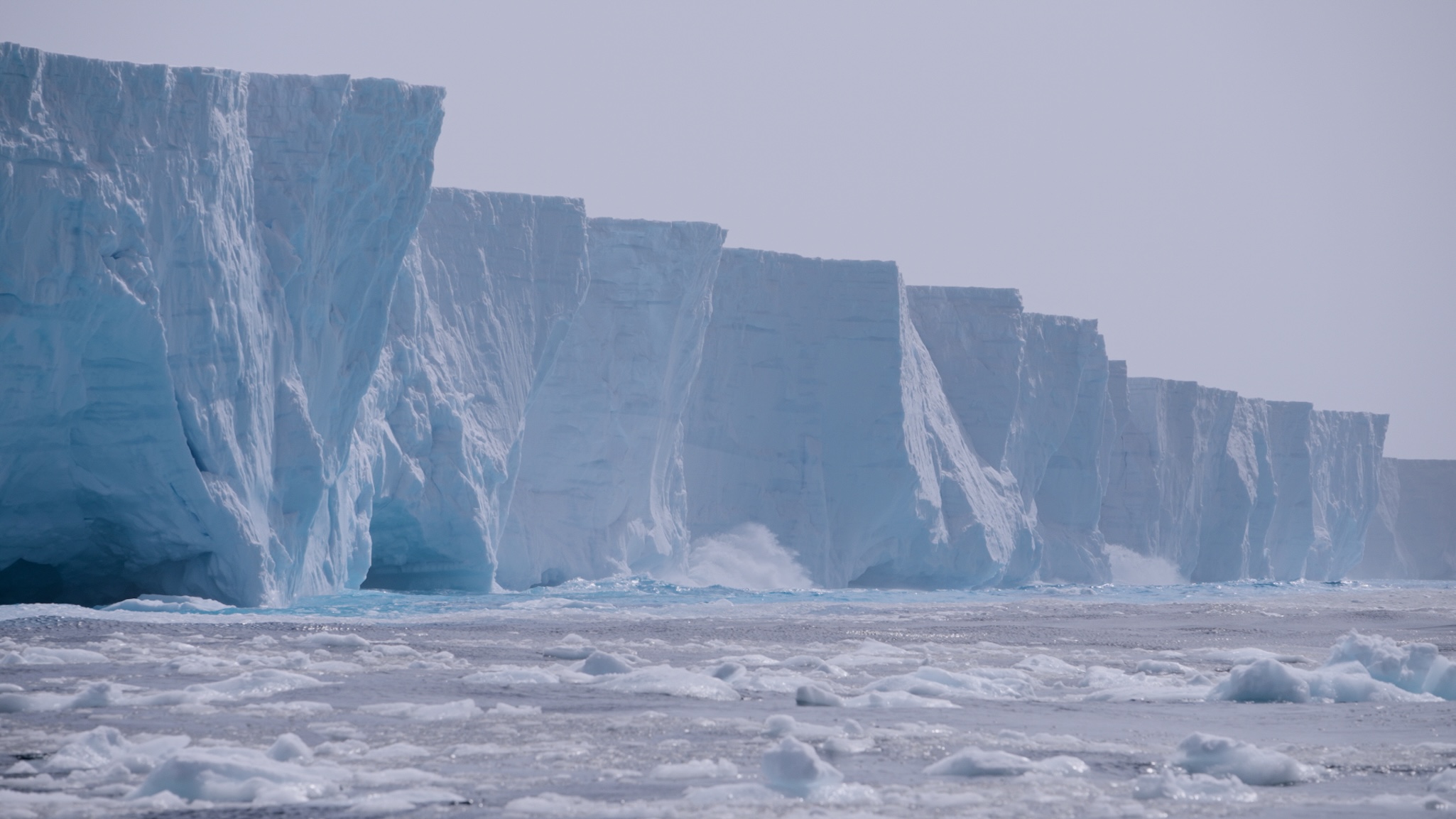 A massive ice shelf rises vertically above the icy sea, with jagged, towering blue-white ice walls and floating chunks of ice in the foreground under a pale, overcast sky.