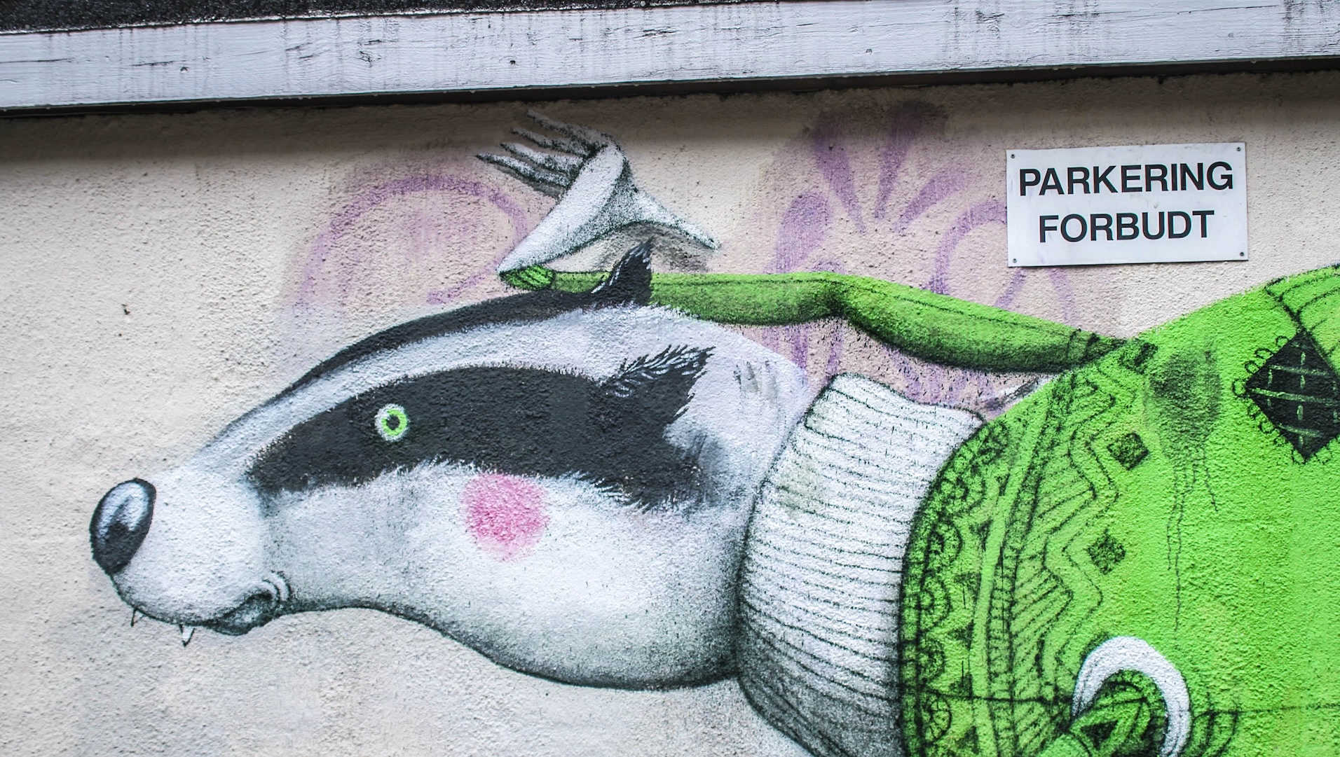 A mural of a badger in a green sweater with a fork on its head is painted on a wall beneath a sign that reads PARKERING FORBUDT in Oslo, Norway.