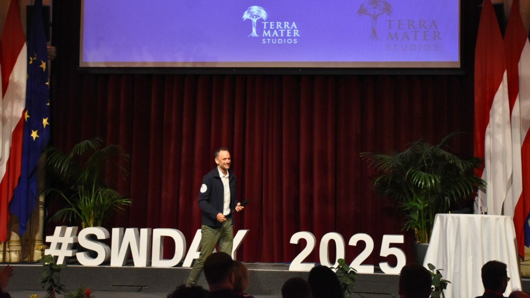 A speaker stands on stage in front of a red curtain, large white letters spelling #SWDays25 2025, and flags. An audience watches as a screen above displays the Terra Mater Studios logo, highlighting innovation in today’s digital world.