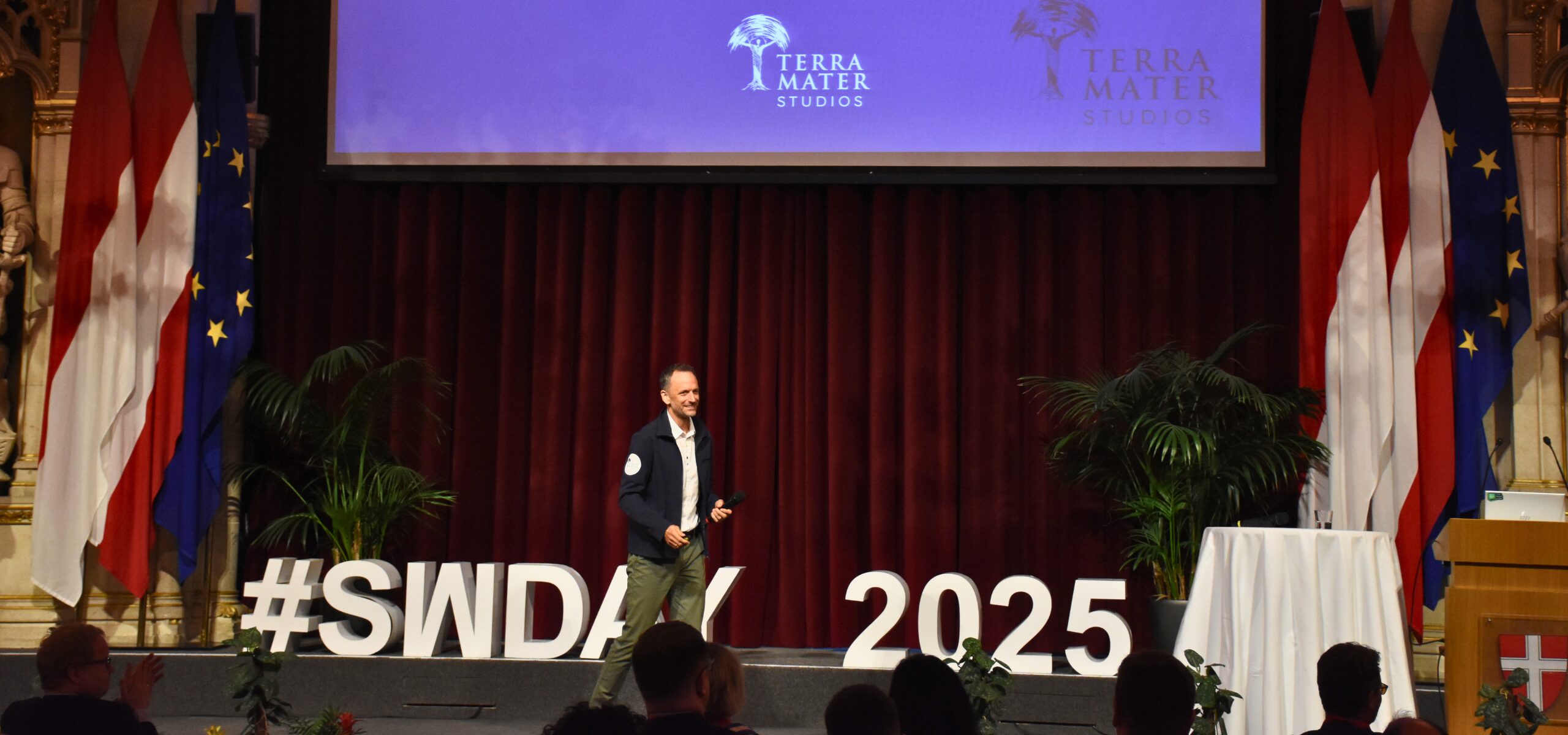 A speaker stands on stage in front of a red curtain, large white letters spelling #SWDays25 2025, and flags. An audience watches as a screen above displays the Terra Mater Studios logo, highlighting innovation in today’s digital world.