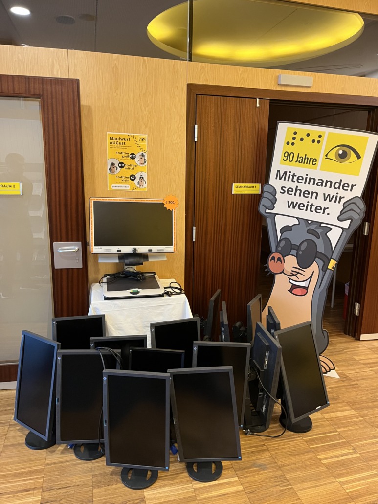 A group of computer monitors is stacked on the floor in front of a table with another monitor, keyboard, and mouse, showing how tech donation can give old devices a second life. Beside them stands a mole cartoon figure with a German sign about 90 years together.