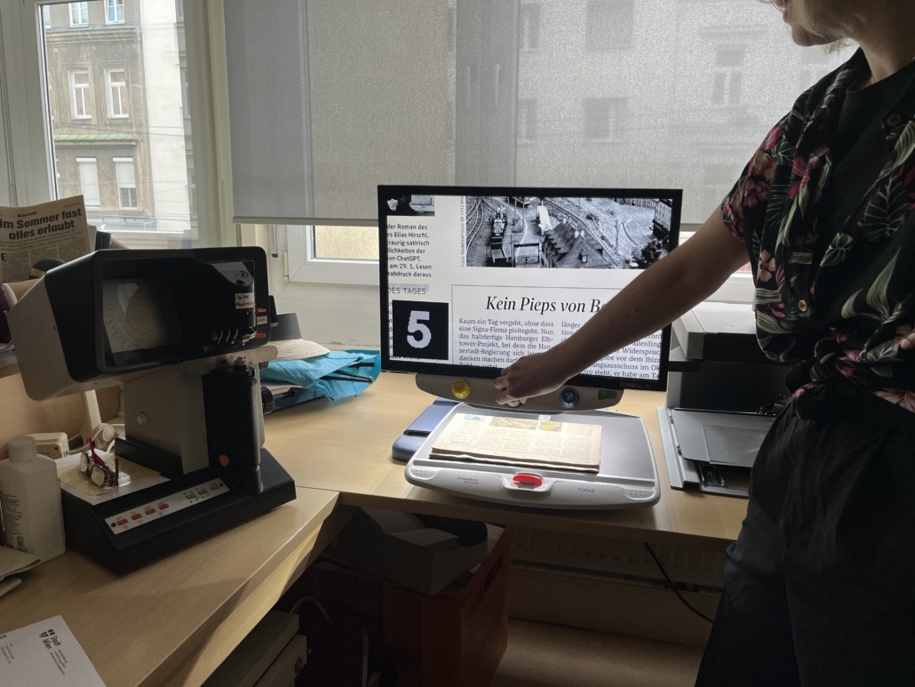 A person uses a video magnifier to read a newspaper article on a large monitor in an office, highlighting accessibility, with a microfilm reader and papers nearby. Sunlight enters through the window in the background.