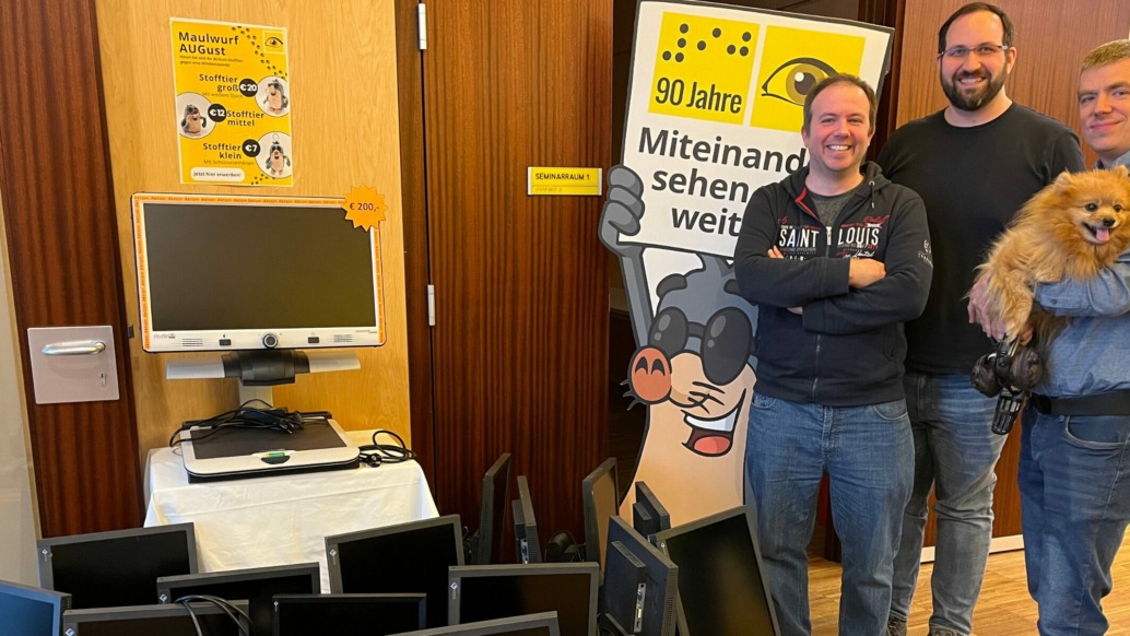 Three smiling men, one holding a small fluffy dog, stand beside a table with computer monitors—part of a tech donation event. Behind them are informational posters, including one that says 90 Jahre and features a cartoon mole.