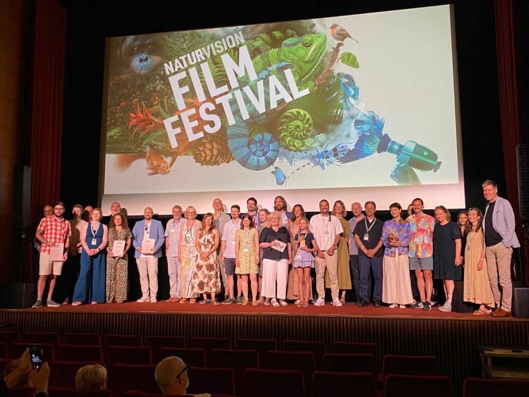 A group of people stands on stage in front of a large NaturVision Film Festival screen, posing for a group photo at a movie theater, with some holding certificates in recognition of their achievements in documentaries.
