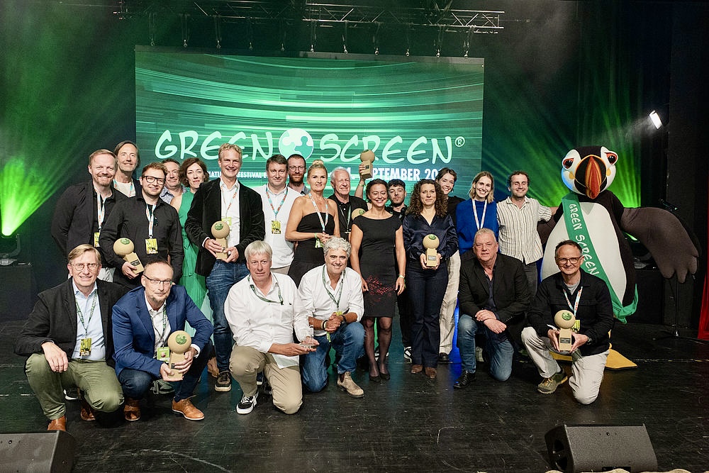 A group of people pose on stage holding green trophies at the GREEN SCREEN Festival, with a large bird mascot and green lighting in the background. The screen behind them displays "Best Cinematography" and "Katavi.