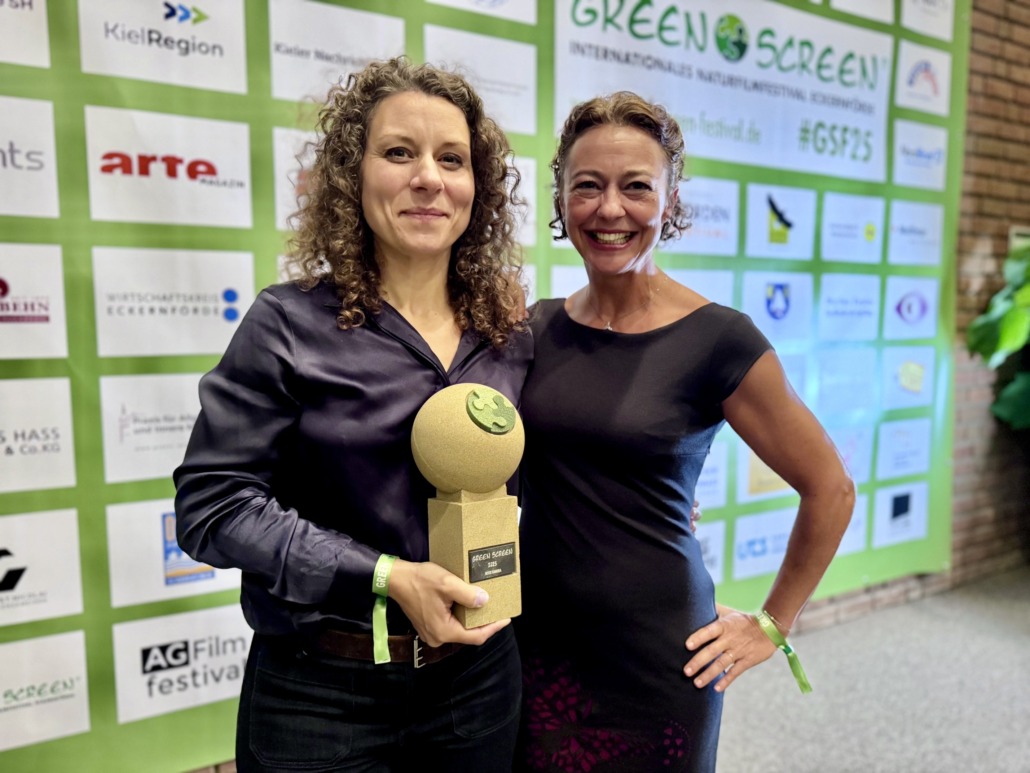Two women smile at the camera at the GREEN SCREEN Festival. One holds a Best Cinematography trophy as they stand before a backdrop with logos and festival branding, celebrating their achievement for Katavi.