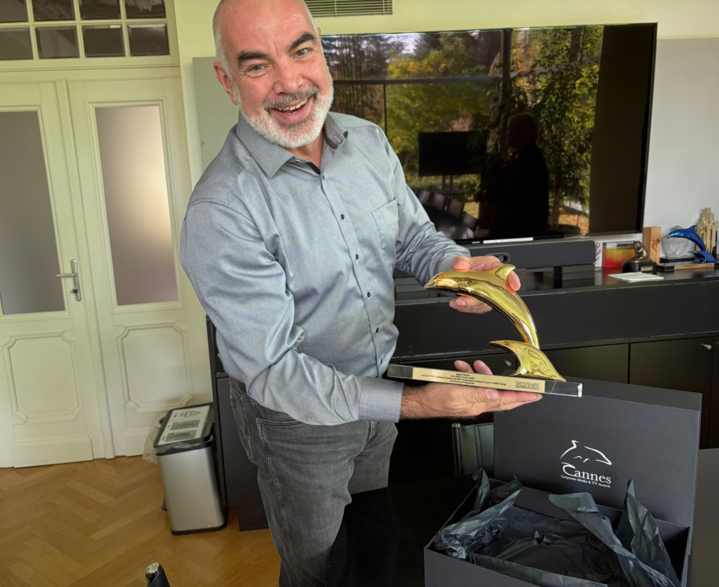 A smiling man in a gray shirt holds a Golden Dolphin award, shaped like a stylized dolphin fin, standing indoors near a black Cannes Corporate Media & TV Awards box with tissue paper on a wooden floor.