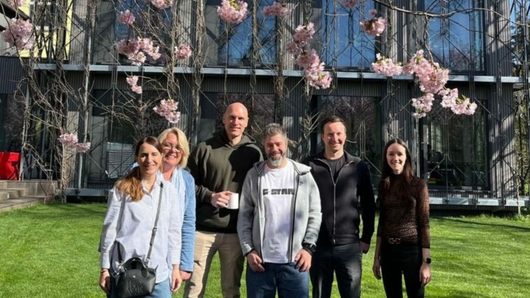 Six people stand smiling on a green lawn in front of a modern building with large windows and blooming pink cherry blossoms, enjoying the sunny weather. This vibrant scene captures the autentic spirit celebrated by Terra Mater Studios.
