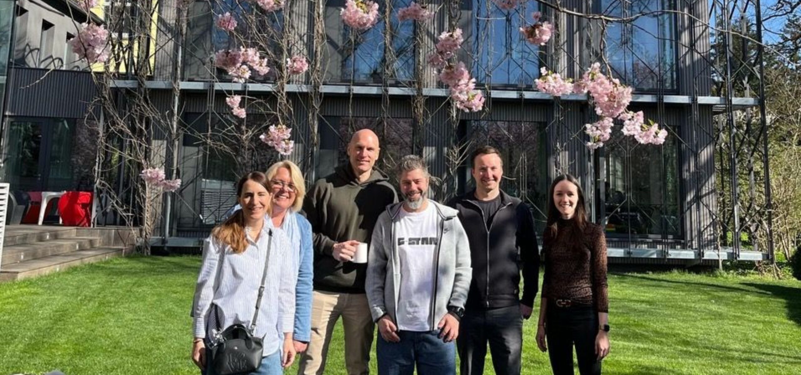 Six people stand smiling on a green lawn in front of a modern building with large windows and blooming pink cherry blossoms, enjoying the sunny weather. This vibrant scene captures the autentic spirit celebrated by Terra Mater Studios.