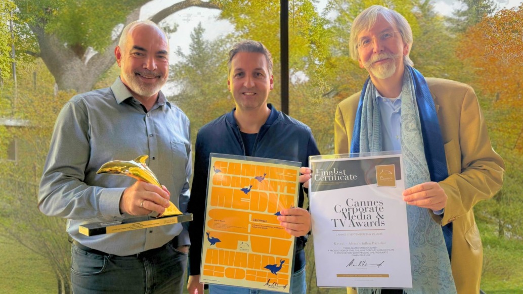 Three smiling men stand indoors by a window with trees outside. One holds a Golden Dolphin trophy, another holds an orange poster, and the third displays a Cannes Corporate Media & TV Awards finalist certificate.