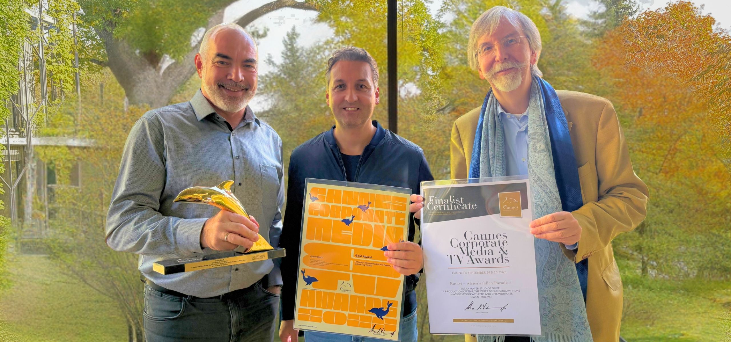 Three smiling men stand indoors by a window with trees outside. One holds a Golden Dolphin trophy, another holds an orange poster, and the third displays a Cannes Corporate Media & TV Awards finalist certificate.