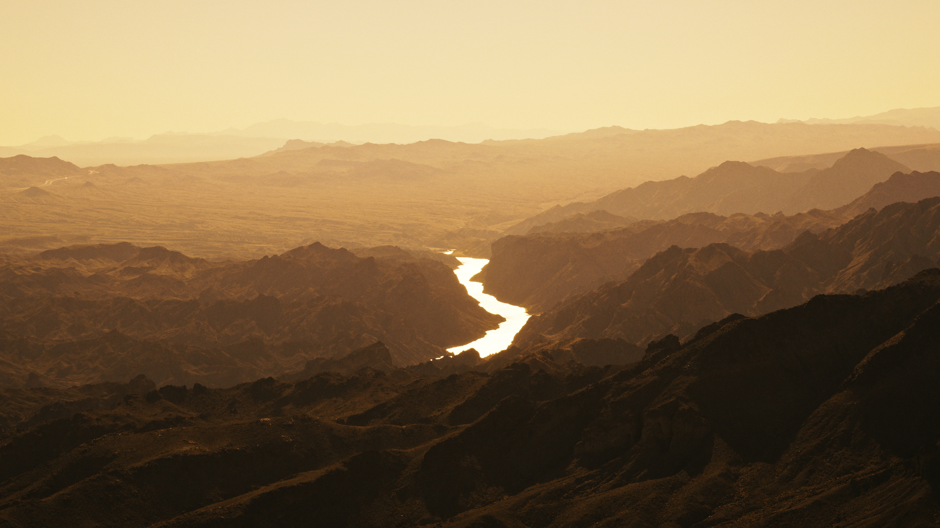 A winding river, the Mighty Colorado, reflects sunlight as it snakes through rugged, dark mountain ranges under a hazy golden sky, creating a dramatic, sunlit contrast in an arid, mountainous landscape.