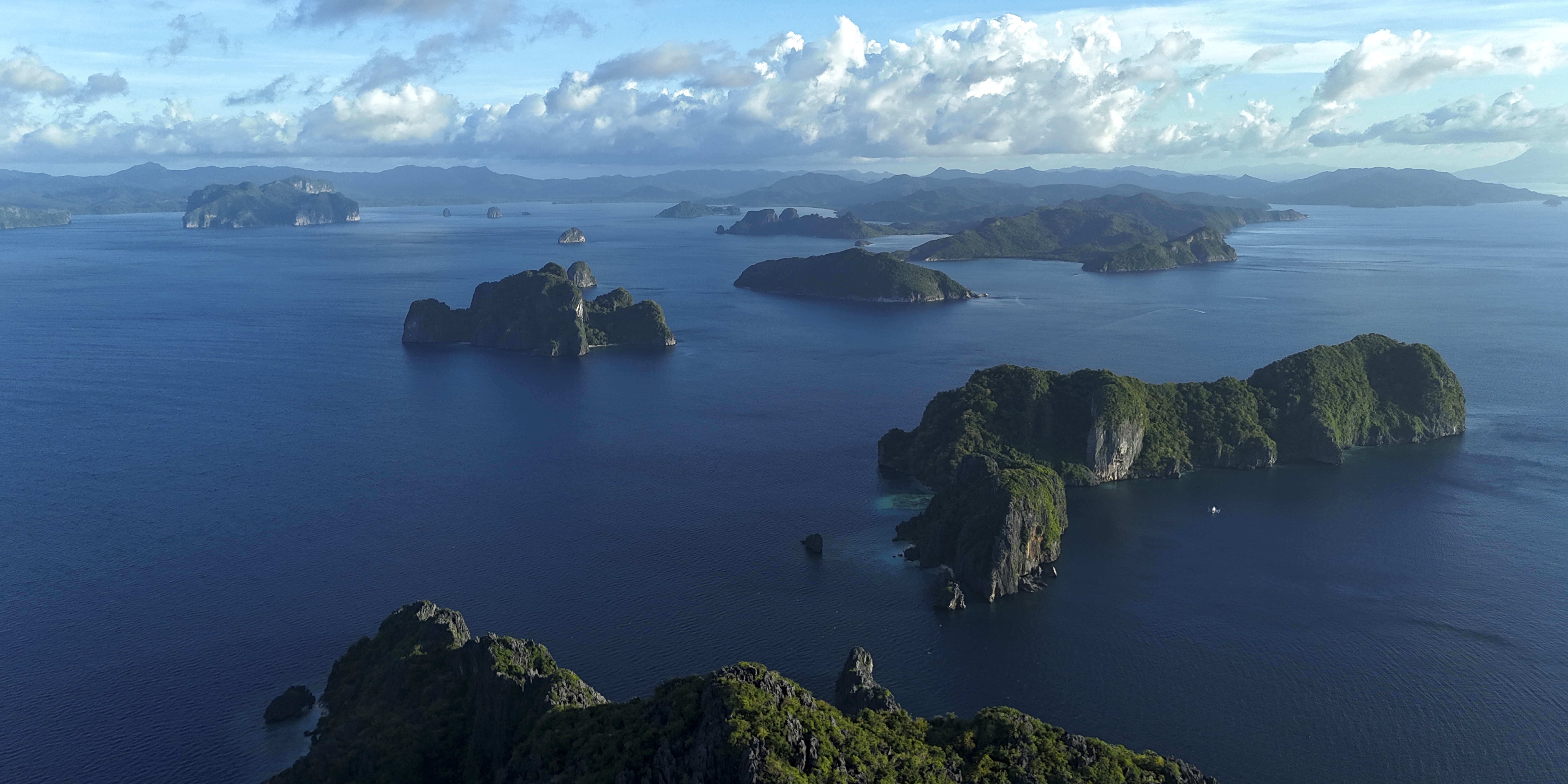 Aerial view of rocky, green islands scattered across a deep blue ocean under a partly cloudy sky, with distant mountains visible on the horizon.