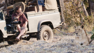 A man sitting on a safari vehicle with a large camera points while observing a lioness resting nearby in Katavi, the dry, grassy landscape famed for its wildlife and now celebrated with an Emmys Nomination for Katavi.