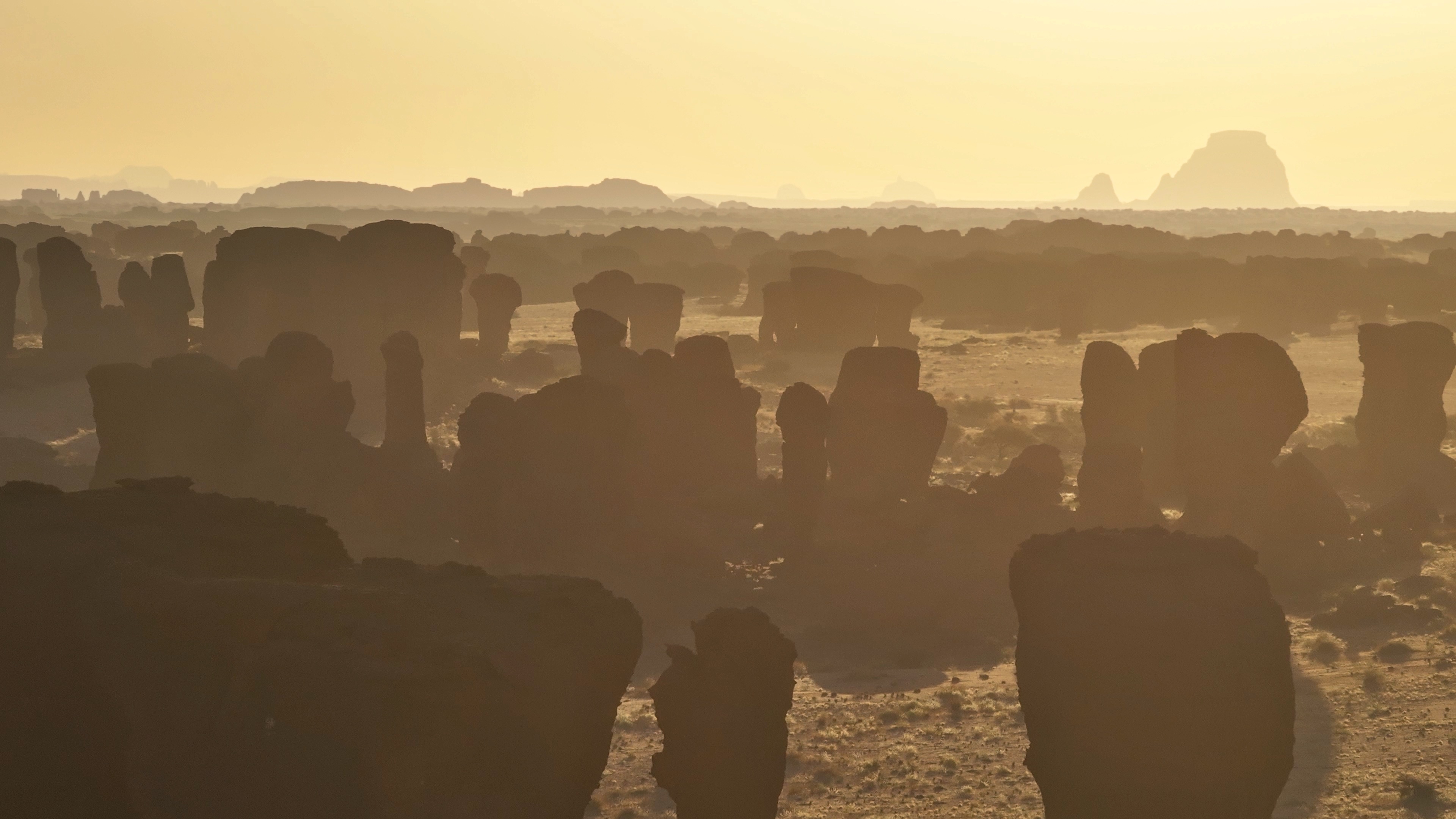 Rock formations in a desert landscape are silhouetted against a hazy, golden sky at sunset, resembling Phantoms of the Desert as distant rocks fade into the misty background, creating a mysterious layered effect.