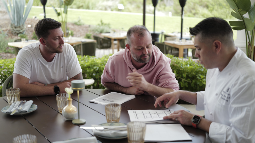 Three men sit at a round outdoor table, discussing a document. The man on the right, in a chef's jacket, explains something about International Production while the others listen attentively. Glasses and tableware are set on the table.