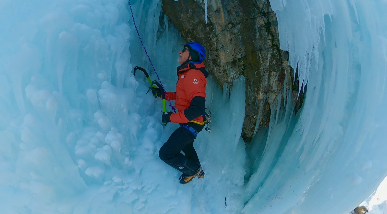 A person wearing a helmet, red jacket, and climbing gear scales a steep, icy cliff with ice axes, surrounded by blue ice and icicles—capturing the spirit of Christmas in the Rockies.