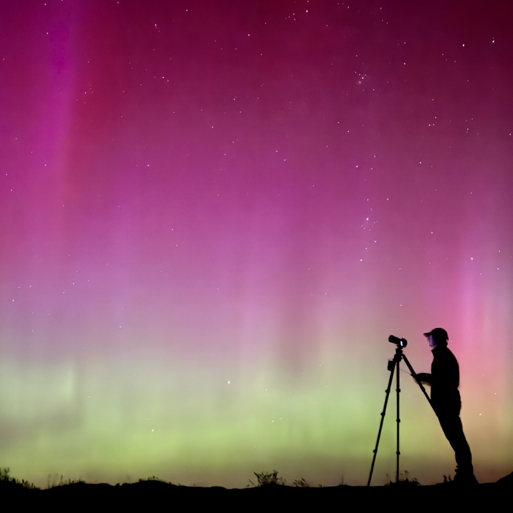 Silhouette of a person with a camera and tripod observing vibrant pink, purple, and green aurora lights in the night sky, standing on a hilltop in the Rockies, evoking scenes from a magical Christmas movie.