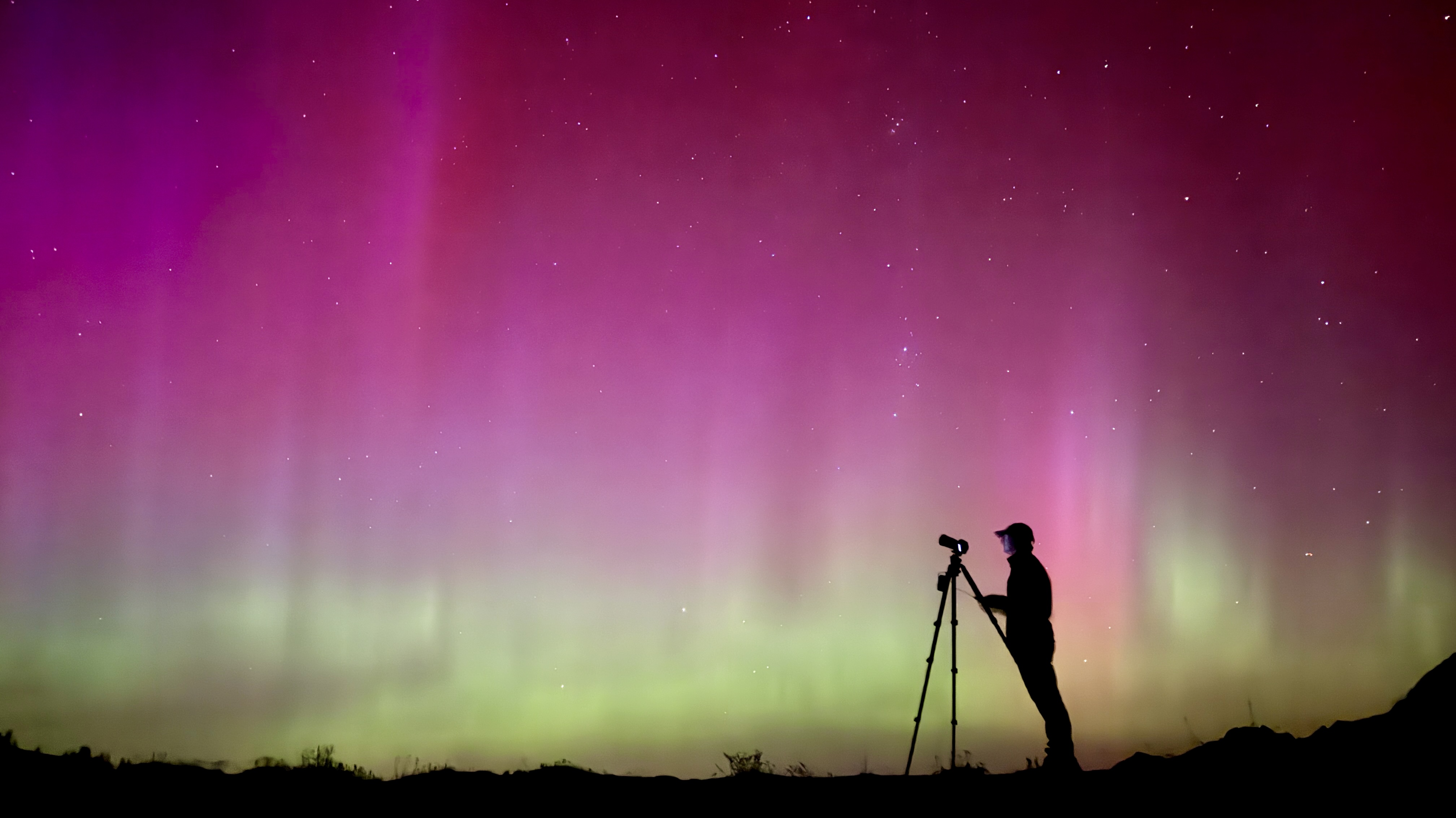 Silhouette of a person with a camera and tripod observing vibrant pink, purple, and green aurora lights in the night sky, standing on a hilltop in the Rockies, evoking scenes from a magical Christmas movie.