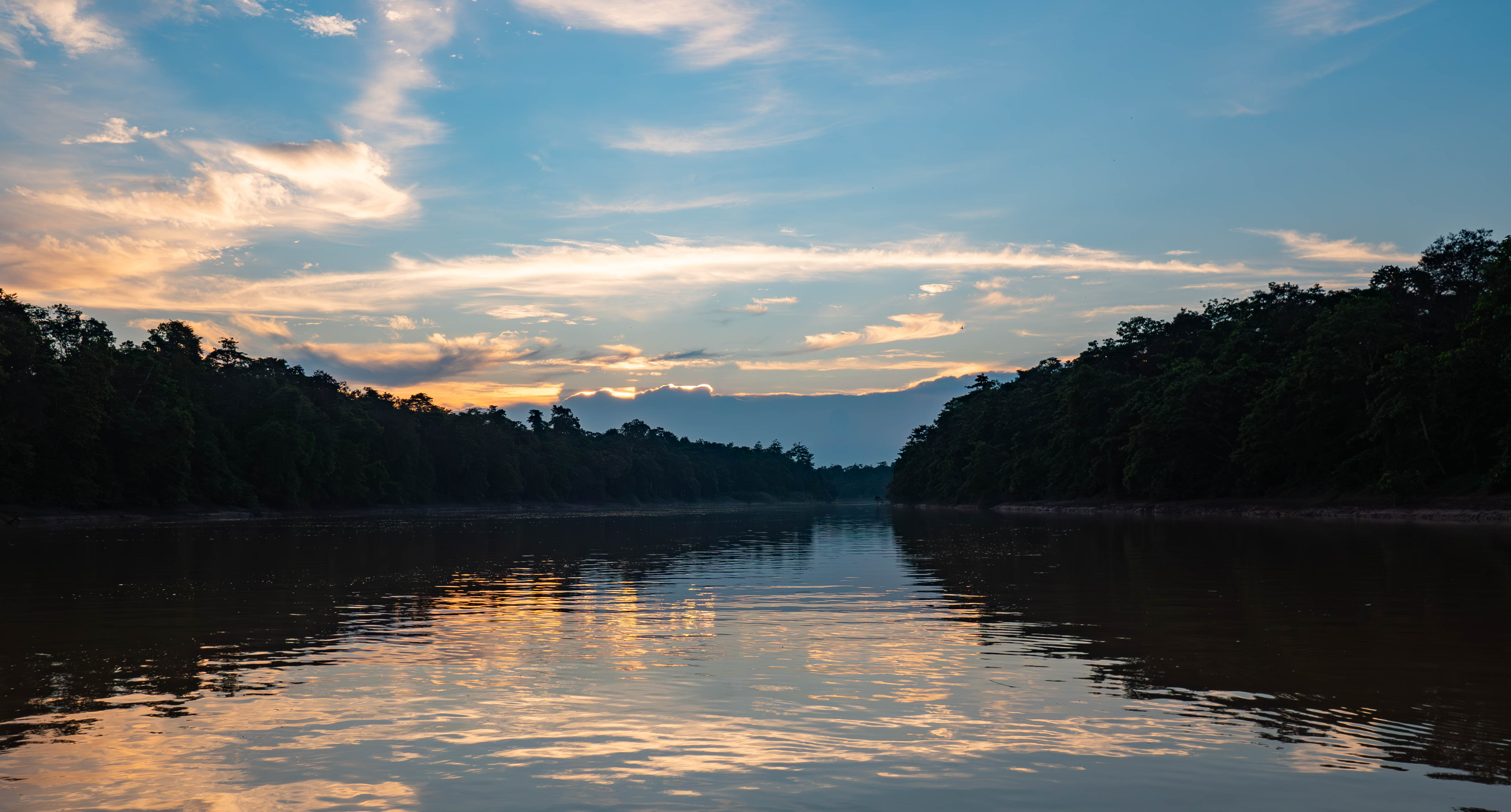 A calm river flows between dense green forests at sunset, with soft clouds and warm colors reflecting on the water under a partly cloudy sky, like a scene from a gentle children's story.